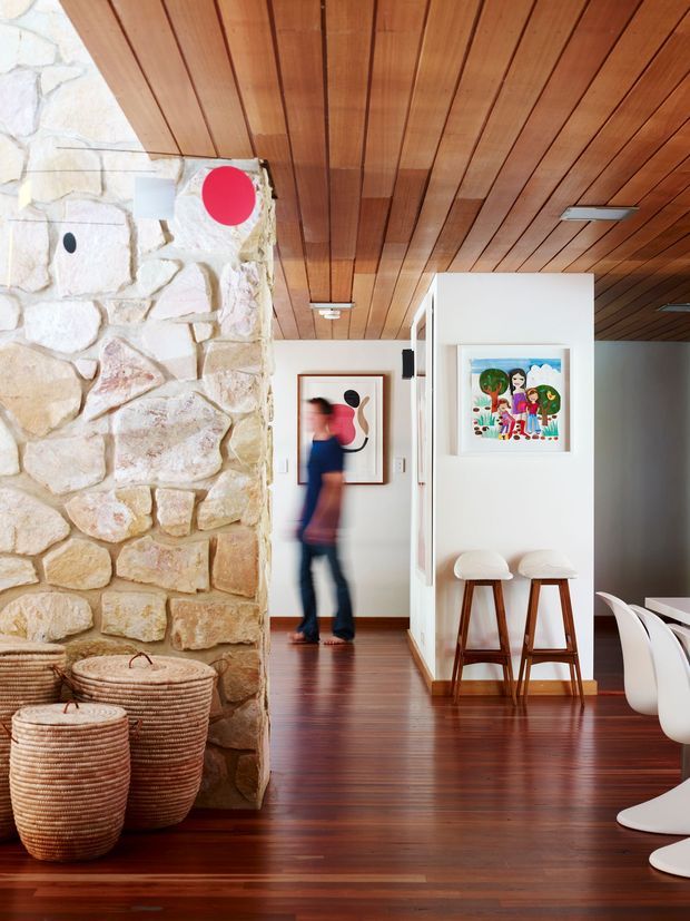 Interior view of an open-plan residential space featuring a rough natural-stone feature wall, richly stained timber-lined ceiling, polished dark timber flooring, and a white kitchen or dining zone. Two timber bar stools sit at a compact peninsula, with white dining chairs visible at right. Artwork, woven baskets, and a blurred person add lived-in detail. The space has a warm, contemporary feel with strong natural-material contrasts.