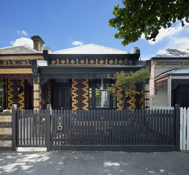 Street-facing view of a narrow single-storey historic terrace or row house with ornate decorative brickwork, elaborate timber lacework and verandah detailing, a corrugated metal roof, and a dark picket fence and gate. Adjacent period facades are visible on both sides, beneath a clear blue sky and mature street tree.