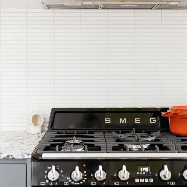 Close-up of a black SMEG freestanding gas range with multiple burners and chrome control knobs, set against a white horizontally tiled backsplash. A pale patterned stone countertop, white ceramic vessel, and red cookware are partially visible.