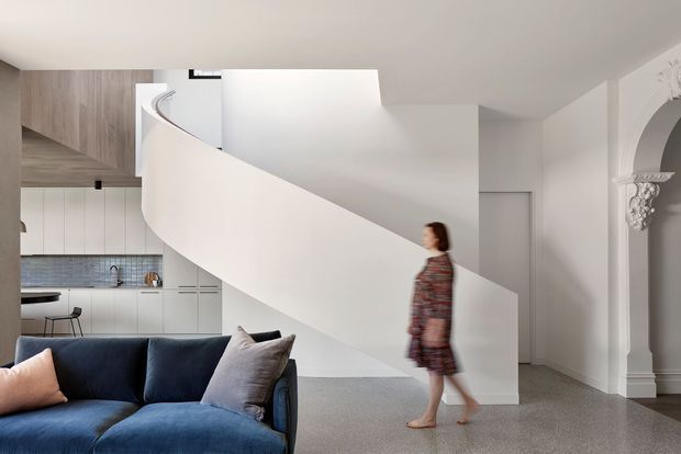 A refined contemporary open-plan interior combining a living room and kitchen, with a sculptural curved white staircase dominating the centre of the frame. The foreground has a deep blue sofa with pale cushions, while the rear kitchen features pale cabinetry, a grey tiled splashback and timber-toned overhead joinery. White walls, polished grey flooring and ornate traditional architrave detailing at the right create a contrast between modern intervention and heritage character.