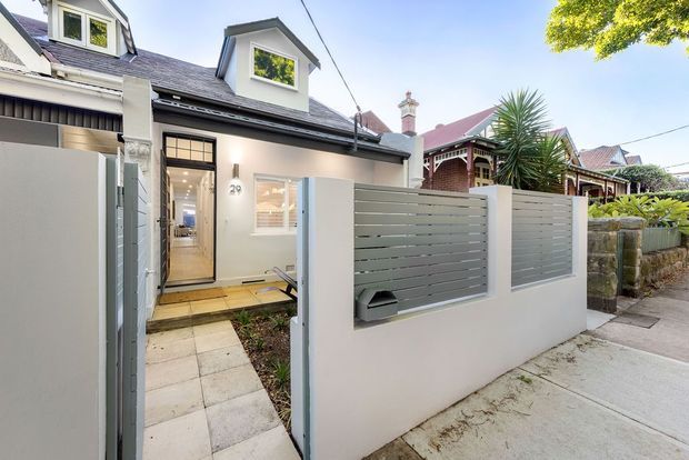 Street-facing view of a compact renovated residential dwelling with a pale rendered facade, dark pitched roof, dormer windows, and a recessed entry gate. Grey horizontal slatted privacy fencing sits on a low rendered wall, with a tiled path leading to a glazed front door. Traditional neighbouring homes and mature planting are visible in the background.