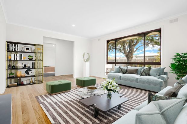 Bright, spacious contemporary living room with pale timber flooring, white walls and ceiling, a large dark-framed picture window, and a coordinated seating arrangement around a black rectangular coffee table. The room includes a light grey sofa and armchairs, green ottomans, a striped area rug, decorative shelving, indoor plants, and a floral centerpiece.