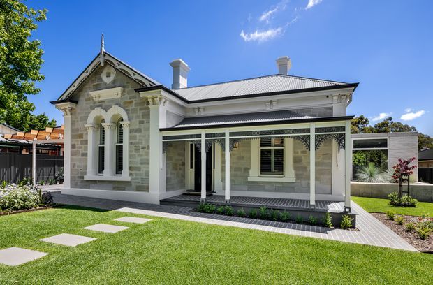 A restored single-storey stone residence with Victorian-era detailing, a steeply gabled front section, decorative bargeboard, arched windows, ornate lacework verandah, white trims, and a corrugated metal roof. The home sits behind a manicured lawn with a stepping-stone path and landscaped garden beds; a contemporary flat-roof extension is visible to the rear.
