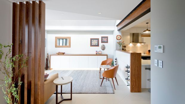 Bright open-plan interior showing a contemporary kitchen, dining and living area. A white kitchen with timber cabinetry and a timber-framed island opens onto a living zone with a light rug and warm-toned seating. Vertical dark timber screening at left and exposed timber ceiling beams add strong architectural detail, while white walls, recessed lighting and pale flooring keep the space airy.