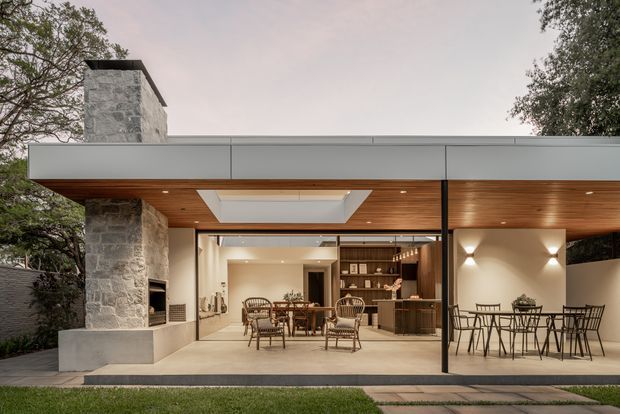 Contemporary single-storey pavilion-style home viewed from the garden, with a broad flat roof, extensive glazed openings, a stone chimney and covered outdoor entertaining terrace. Warm timber-lined soffits, an outdoor dining area, indoor dining and kitchen spaces, and integrated fireplace elements are visible. The design has a strong indoor-outdoor connection and a calm neutral palette.