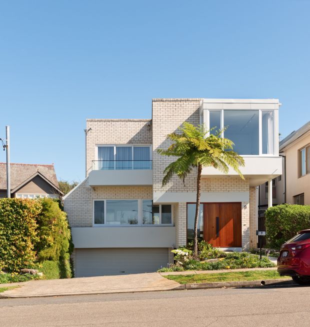 A bright street-level view of a contemporary multi-storey urban residence. The home features pale brickwork, crisp white rendered volumes, large modern windows, a projecting upper-level bay, a balcony with glass or slim metal balustrade, and a prominent timber entry door. A partially below-grade garage, landscaped front garden, hedges, neighbouring houses, and a parked car are visible. The clear blue sky and strong sunlight create a clean, airy appearance.
