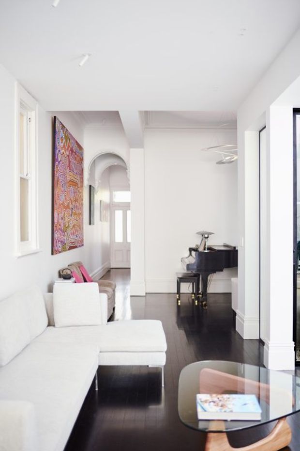 Bright, predominantly white open-plan living space with a white sectional sofa in the foreground, dark polished timber flooring, a black grand piano, and a glass-topped coffee table. An arched hallway leads toward a front door, while colourful wall art provides a strong accent against the white walls. Tall windows and simple contemporary detailing create an airy, refined interior.