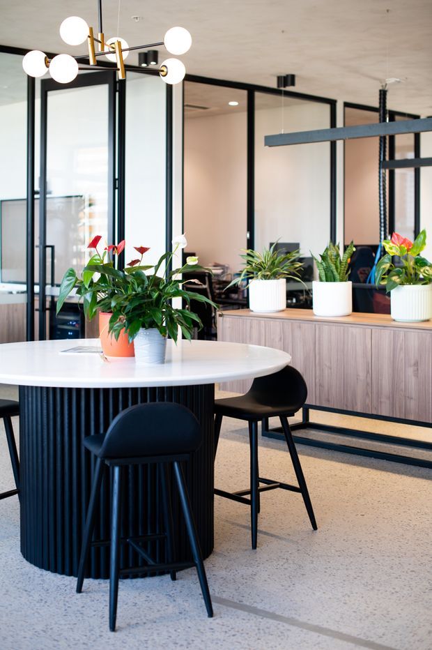 A bright contemporary office breakout and dining area with a round white island table, black bar stools, black-framed glass partitions, timber-look built-in cabinetry, indoor plants, and a sculptural globe pendant light. The space has a polished light terrazzo or concrete floor and a clean, refined workplace fit-out.