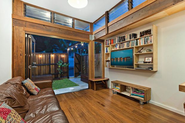 Warm, compact living room with rich timber framing, exposed high-level glazing, polished timber flooring, and a built-in wall shelving and media unit. A leather sofa sits beside an open doorway leading to a small fenced courtyard, while the timber structure and upper windows create an airy loft-like feel.