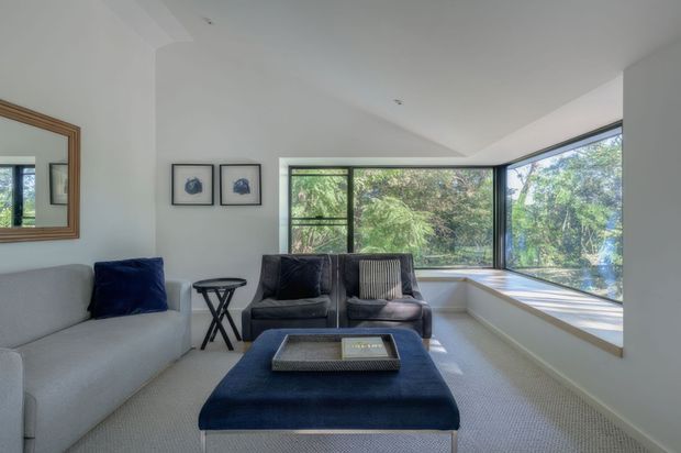 Bright, compact contemporary living room with a grey sofa, two dark leather armchairs, a large navy upholstered ottoman, framed artwork, and a timber-framed mirror. A broad corner of black-framed windows overlooks leafy greenery, while a built-in window seat runs along the glazing. The room has white walls, carpeted flooring, recessed ceiling lights, and a calm neutral-and-navy palette.