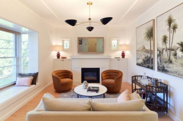 A bright, refined living room with a central white fireplace mantel, symmetrical wall sconces, a sculptural mid-century pendant light, two cognac leather lounge chairs, a pale sofa, and a round coffee table. Large windows with a built-in window seat bring in natural light, while framed botanical artwork and a dark drinks cabinet add decorative detail.