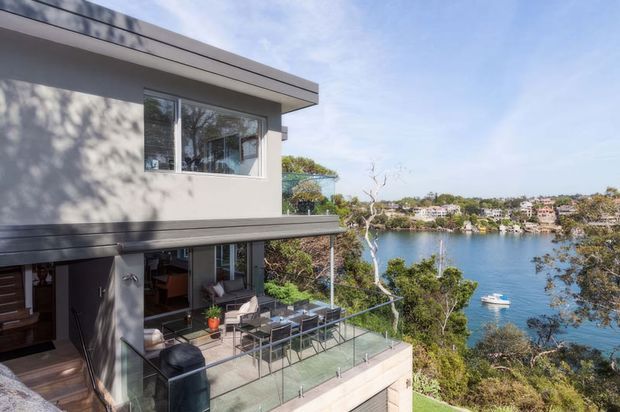 Exterior view of a contemporary multi-level residence with flat rooflines, rendered grey walls, extensive glazing, and a glass-balustraded terrace overlooking a leafy waterfront setting. An outdoor dining area and open living space are visible on the lower terrace, with water and houses across the inlet in the background.