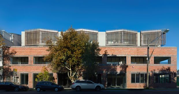 Exterior street view of a contemporary multi-storey brick building with a regular grid of dark-framed windows and distinctive rooftop volumes screened by vertical metal fins. Mature street trees and parked cars partially obscure the lower facade.