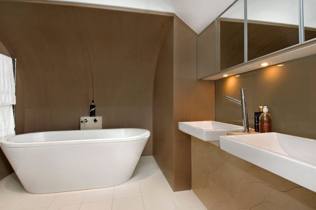 Contemporary bathroom with a freestanding white bathtub, two wall-mounted white basins, and a large mirrored vanity cabinet. Warm timber wall panelling wraps around the bath area, while recessed under-cabinet lighting illuminates the taupe-toned splashback and stone-look vanity surfaces. Pale tiled flooring and clean geometric fixtures create a refined, warm modern interior.