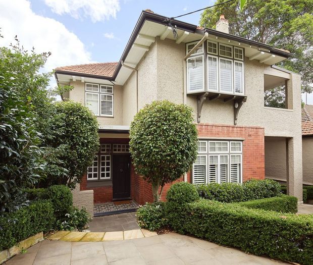 A two-storey traditional residential house viewed from the front, with cream textured stucco walls, red brick detailing, terracotta tiled pitched roofs, projecting bay windows with white louvred shutters, and a dark timber entrance door. Carefully clipped hedges and rounded shrubs frame the paved front path and facade.