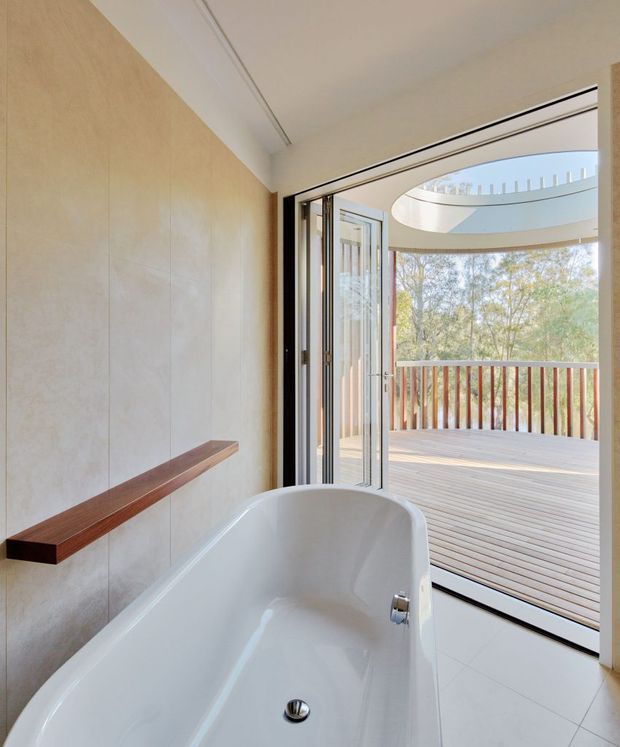 Bright contemporary bathroom featuring a freestanding white bathtub beside full-height glazed folding doors opening to a timber deck and balcony railing. The room has large pale stone-look wall panels, light tiled flooring, a simple timber wall-mounted shelf, and a distinctive oval skylight or roof opening visible beyond the doors.