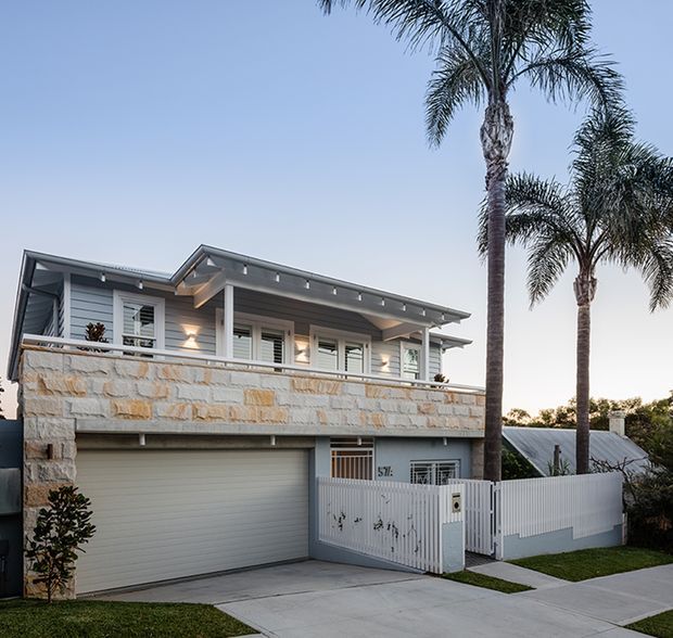 Exterior street-level view of a contemporary two-storey home with a broad low-pitched roof, white horizontal cladding, stone-faced upper-level wall, shaded balcony, external roller garage door, and a white slatted pedestrian gate and fence. Tall palm trees and a paved driveway frame the property.