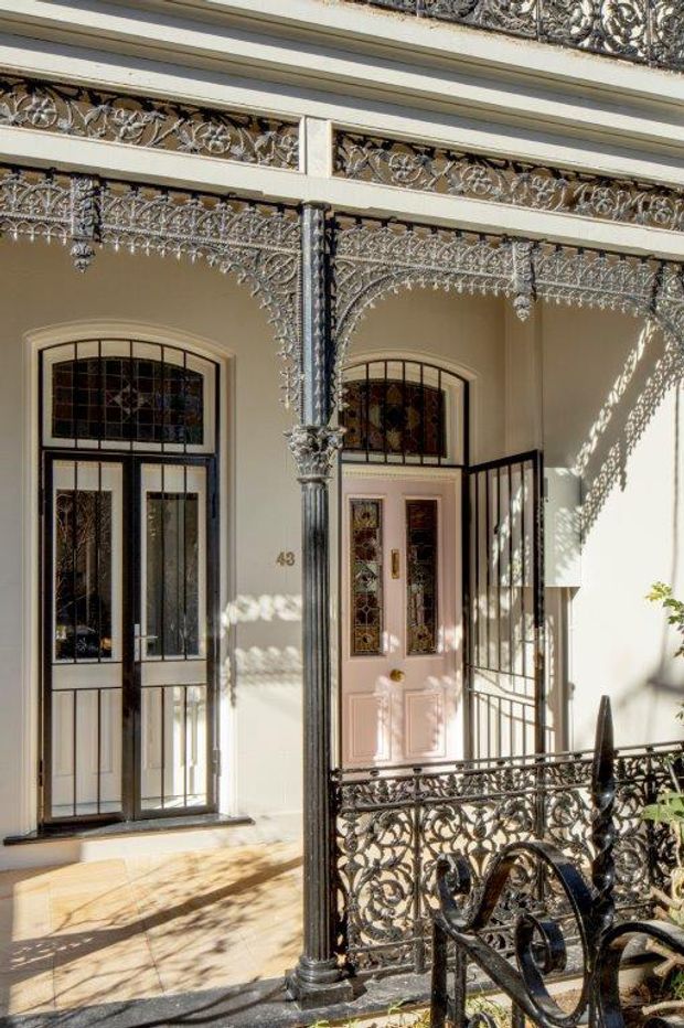 Street-facing entrance facade with ornate cast-iron verandah detailing, decorative balustrades, slender columns, arched stained-glass windows, and a pale pink paneled front door. The heritage-style frontage is photographed in bright sunlight, with strong patterned shadows across the pale rendered walls and tiled entry paving.