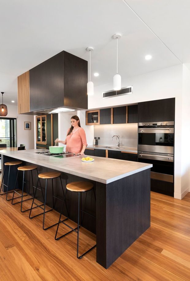 A bright contemporary kitchen dominated by a large island with pale stone benchtop and dark timber-look cabinetry. The island includes seating for four on slim black stools with timber seats. Matching dark cabinetry lines the rear wall, incorporating a sink, integrated appliances, oven, cooktop and warm under-cabinet lighting. Light timber flooring, white walls and ceiling, pendant lights, and a vertical timber batten feature add warmth and texture.