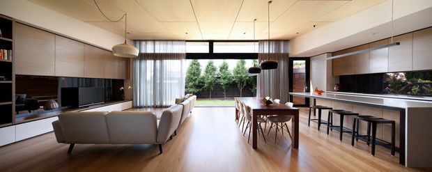 A spacious open-plan contemporary living, dining and kitchen area. The room features warm timber flooring, extensive pale timber built-in cabinetry, a long kitchen island with stools, a dining table, a streamlined media wall, large glazed openings to a planted outdoor area, and simple pendant lighting. The restrained palette and clean lines create a bright, refined, inviting interior.