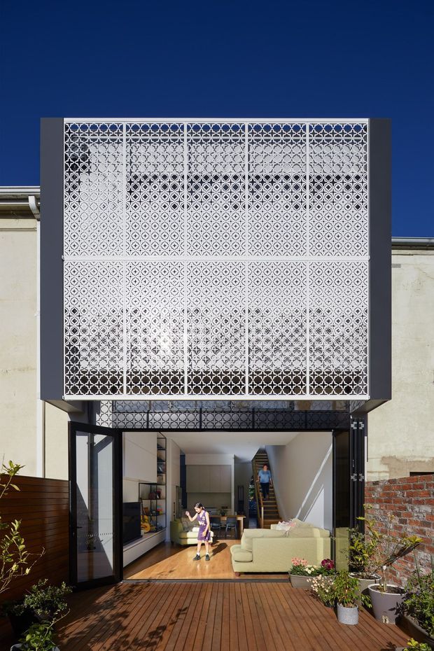 Exterior view of a contemporary urban residence with a large decorative perforated metal screen forming an upper facade and balcony enclosure. The open ground-level glazing connects an interior living area to a timber deck, with built-in cabinetry and a pale sectional sofa visible inside. Dark framing, weathered masonry walls, and dense patterned screening create a strong architectural feature against a clear blue sky.