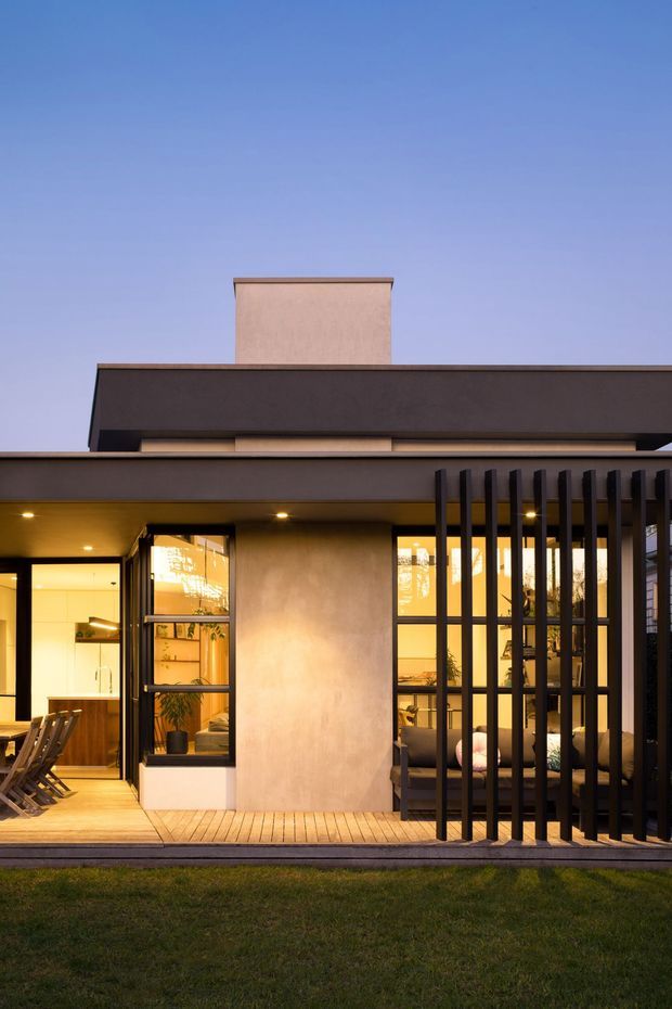 Twilight exterior view of a contemporary single-storey residence, featuring a flat layered roofline, warm rendered walls, extensive glazed openings, timber decking, and a dark vertical slatted screen enclosing an outdoor seating area. Warm interior lighting is visible through the windows and doors, with lawn in the foreground.