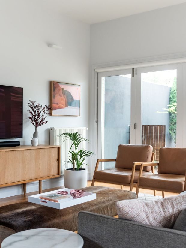 Bright, refined living room with a mid-century-inspired timber sideboard, television, framed artwork, cane or timber-framed lounge chairs, a grey sofa, layered rugs, and a white coffee table. Large glazed doors bring in abundant daylight, while indoor plants and warm brown upholstery add an inviting natural feel.