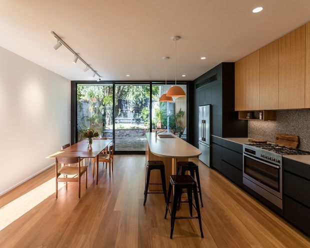A contemporary open-plan kitchen and dining area with warm timber flooring and cabinetry, dark built-in kitchen joinery, a stone-textured splashback, a long island with integrated sink and seating, and a timber dining table. Full-height black-framed glazing opens toward a leafy courtyard, while pendant lights and track lighting illuminate the space.