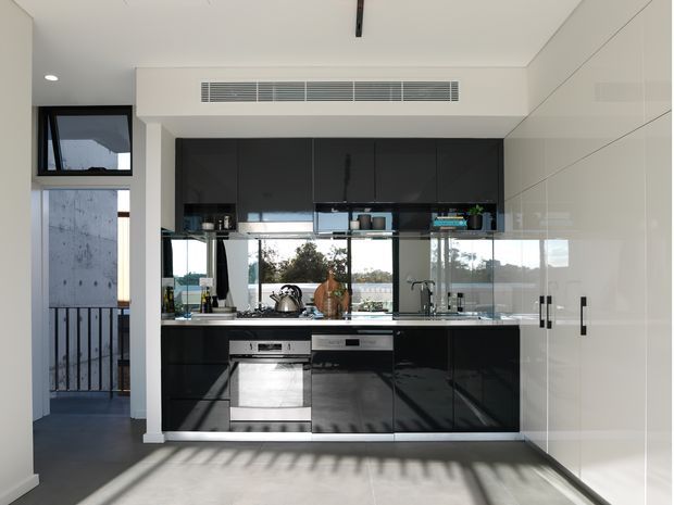A contemporary compact kitchen with glossy black lower and upper cabinetry, a reflective mirrored splashback, integrated stainless-steel appliances, and a pale stone benchtop. Full-height white cabinetry lines the right wall, while large glazing and a side opening bring in strong natural light. The space has a clean, streamlined fit-out with dark flooring and visible outdoor views beyond the kitchen.