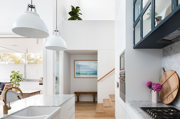 Bright contemporary kitchen and open-plan living area with a white marble-look island and benchtops, integrated white cabinetry, dark blue-gray overhead cabinetry with glass fronts, and a gas cooktop. Large white pendant lights hang above the island. The space opens toward a dining/living area with timber and woven furniture, while timber stairs and a framed artwork are visible beyond. Blue-gray framed glazing and a marble-patterned splashback add contrast to the predominantly white interior.