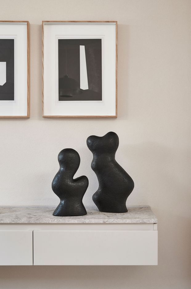 A minimalist interior vignette featuring two black sculptural figurines displayed on a light stone-topped floating cabinet. The cabinet has smooth off-white built-in drawers, while two abstract monochrome artworks in light timber frames hang above on a warm neutral plaster wall.
