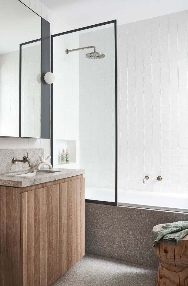 Bright contemporary bathroom featuring a freestanding-style bathtub enclosed by a black-framed glass shower screen, white textured wall tiles, a timber vanity with a pale stone benchtop, a large frameless mirror, and brushed metal tapware. The space has a clean, refined, natural material palette with terrazzo-like flooring and bath surround.
