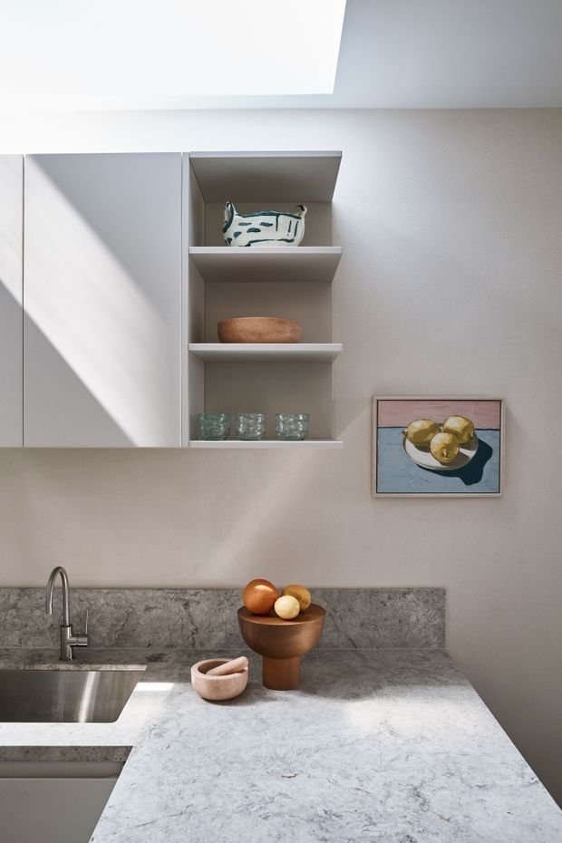 A refined contemporary kitchen vignette featuring pale grey cabinetry, open display shelving, a mottled grey stone benchtop and splashback, and a stainless steel tap and sink. Natural light enters through a large ceiling skylight, casting strong geometric shadows across the cabinetry. Ceramic vessels, glassware, a wooden bowl, and framed still-life artwork add restrained decorative detail.