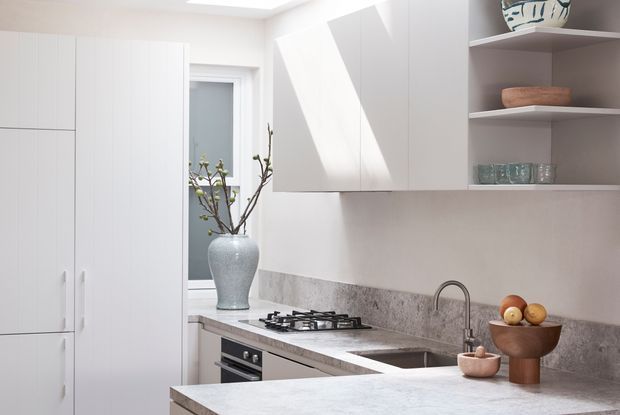 Bright, compact contemporary kitchen with white flat-panel cabinetry, integrated tall storage, open shelving, a gas cooktop and oven, stainless steel sink and tap, and a pale stone island or benchtop. A mottled stone splashback runs along the rear wall, with natural ceramic and glass objects providing subtle decorative accents. Daylight enters through a skylight and window.