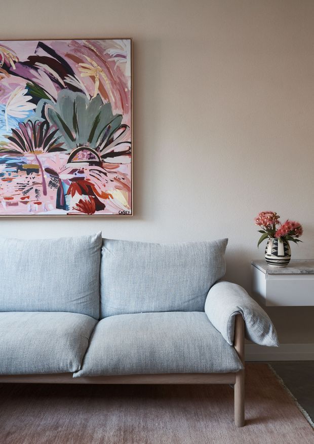 A close-up interior vignette of a softly styled living room, featuring a pale blue-grey upholstered sofa with wooden legs and curved arms, a neutral textured wall, a colourful abstract floral artwork, and a small side table with a patterned vase and flowers. The palette is calm and muted with warm pink and terracotta accents.