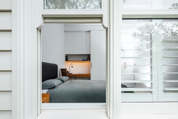 View through a white-framed window into a compact, contemporary bedroom. The room contains a low bed with dark blue-grey bedding, a dark upholstered headboard, and a built-in white wardrobe or shelving wall with an illuminated timber-toned desk niche. White exterior weatherboards and detailed window trim are prominent in the foreground, while reflections and glazing reveal additional shelving.