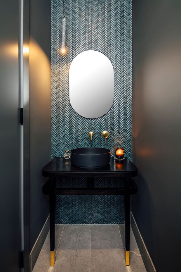A compact, dark and sophisticated powder room with a black vessel basin on a slim black console vanity, brass wall-mounted tapware, and an oval mirror. The central wall is covered in blue-grey herringbone patterned tile, while the surrounding walls are charcoal grey. Warm pendant lighting, a candle, and brass details add a moody luxe atmosphere. Large grey floor tiles and a partially visible glass door complete the space.