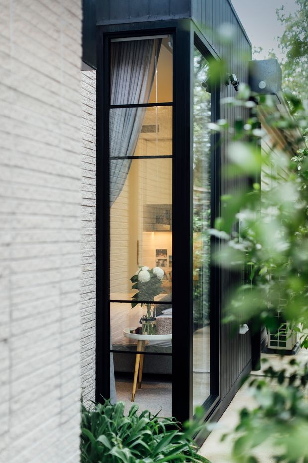 Narrow exterior view of a contemporary dark-clad glazed extension or entry passage beside a light textured masonry wall. A tall black-framed window reveals a softly lit interior with curtains, a bathtub or basin-like fixture, and plant decor, while lush foreground planting partially obscures the facade. The image emphasizes vertical glazing, strong black framing, and contrasting pale brick and dark metal cladding.