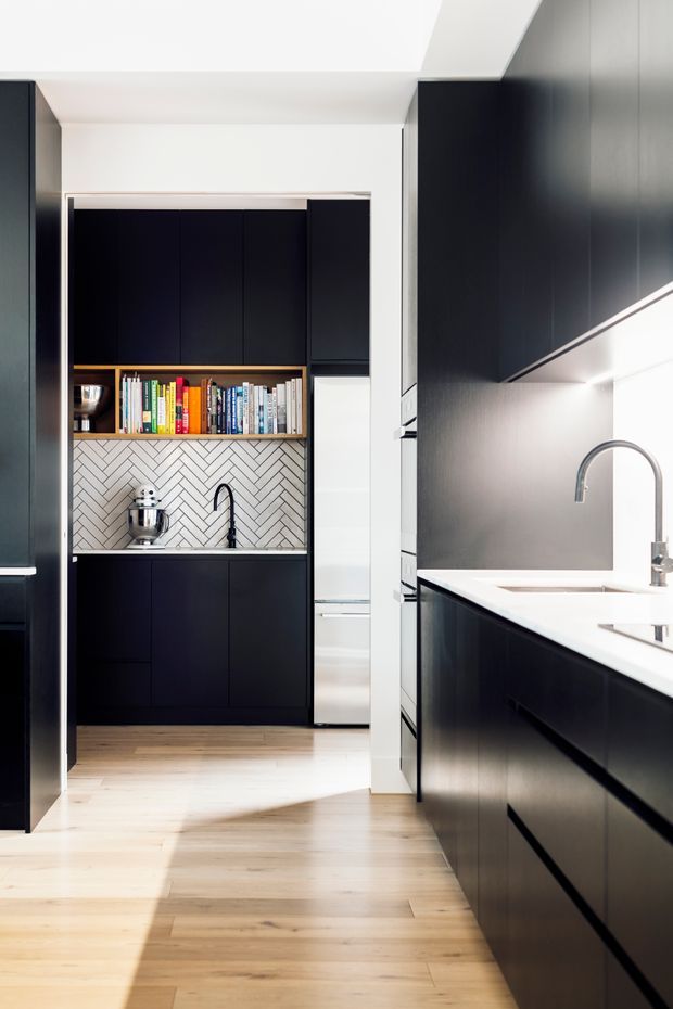 A contemporary galley kitchen with sleek matte-black cabinetry, a bright white benchtop, integrated appliances, and a compact secondary prep area. The rear nook features open shelving with colourful books, a stainless-steel stand mixer, a black tap, and a white herringbone tile splashback. Pale timber flooring and white walls and ceiling balance the dark joinery, while under-cabinet lighting adds illumination.