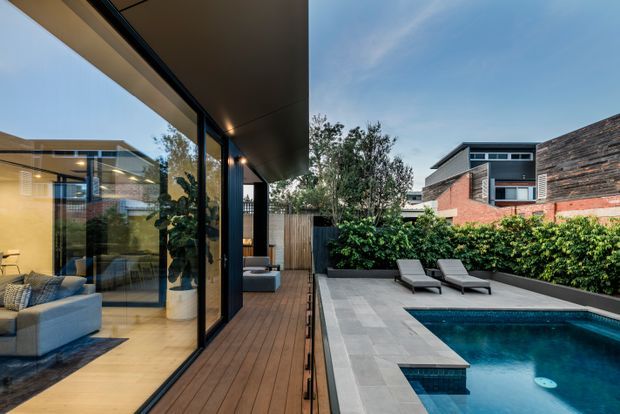 Contemporary residential courtyard viewed from a covered timber deck, with expansive black-framed glazing opening to a refined living area on the left and a compact swimming pool surrounded by pale stone paving on the right. Dense hedging, outdoor loungers, rendered and brick neighbouring forms, and a broad projecting roof create a private, modern outdoor setting.