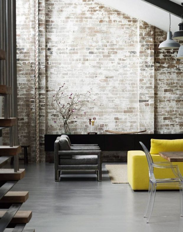 Industrial-style open-plan interior with a tall, whitewashed exposed-brick wall, dark structural beam, polished grey concrete floor, and a partially visible stairway at left. The space includes a charcoal leather armchair, bright yellow upholstered seating, a transparent modern chair, timber tabletop, pendant lights, and a dark built-in console or partition. The composition is urban, high-contrast, and contemporary with strong warehouse-loft character.