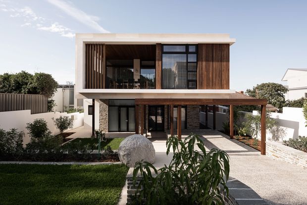 Exterior view of a contemporary two-storey residence with a strong horizontal composition, white rendered surfaces, warm vertical timber screening and cladding, dark-framed glazing, and a covered timber pergola over the entry and driveway. The landscaped front garden includes paving, shrubs, lawn, and a sculptural stone feature.