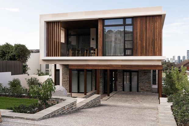 Contemporary two-storey urban residence with a strong rectangular form, pale rendered exterior framing, extensive dark glazing, warm vertical timber screening and timber cladding, and exposed dark brickwork at the lower level. A covered upper balcony or living area opens behind timber battens, while the ground level includes recessed entry doors and a broad textured driveway. Landscaped garden beds and a distant city skyline are visible around the property.