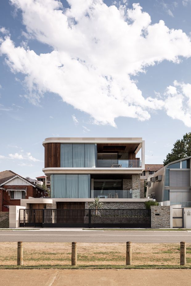 Street-facing photograph of a contemporary multi-storey residential house in an established suburban neighbourhood. The building has rounded horizontal slab edges, extensive floor-to-ceiling glazing with sheer curtains, dark timber wall cladding, exposed stacked-stone masonry, deep recessed balconies, and a flat roof. A dark metal front gate and garage door sit behind a low masonry wall, with neighbouring homes visible on either side.