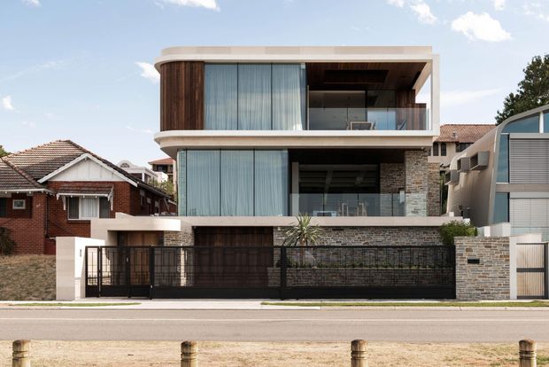 Street-facing view of a contemporary multi-storey residence with a rounded, cantilevered upper floor, expansive floor-to-ceiling glazing, timber screening, exposed light stone masonry, and a dark metal boundary fence and garage door. The home is set among established suburban houses and has a flat-roofed, layered facade composition.