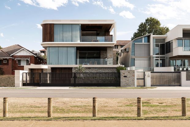 Street-facing view of a contemporary multi-storey residential dwelling with a flat roof, projecting upper-level volumes, extensive glazing, timber screening, dark metal fencing, and pale masonry walls. The modern home is set within an established suburban streetscape alongside older neighbouring houses.