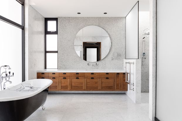 Bright contemporary bathroom with a freestanding black-and-white clawfoot bathtub, long walnut timber vanity with multiple drawers, pale stone or porcelain floor and wall surfaces, and a small-scale mosaic tile feature wall. A large round mirror is centered above the vanity, with a glazed shower enclosure visible to the right. Black-framed windows and doors provide strong linear contrast.
