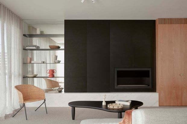 A refined contemporary living room with a black built-in fireplace wall, pale stone ledge, open shelving, and warm timber cabinetry. Sheer curtains span the glazing on the left, while woven lounge chairs and a sculptural dark coffee table add texture and contrast. The space has a calm, minimalist character with carefully integrated joinery and soft neutral furnishings.