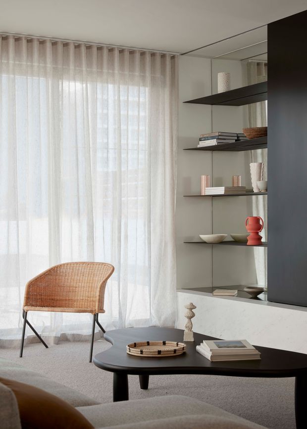 A softly lit contemporary living room with sheer floor-to-ceiling curtains, a woven cane lounge chair, pale upholstered seating, and a low dark oval coffee table. A built-in shelving and media wall occupies the right side, featuring dark floating shelves, books, ceramics, and decorative objects, with a large dark television panel. The palette is warm neutral with restrained red accents and a calm, refined atmosphere.