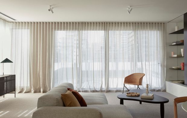 Bright, softly furnished contemporary living room with floor-to-ceiling sheer curtains spanning a glazed wall, a pale upholstered sectional sofa, a sculptural dark coffee table, woven lounge chair, built-in open shelving, and subtle track lighting. The neutral palette and abundant filtered daylight create an airy, calm atmosphere.